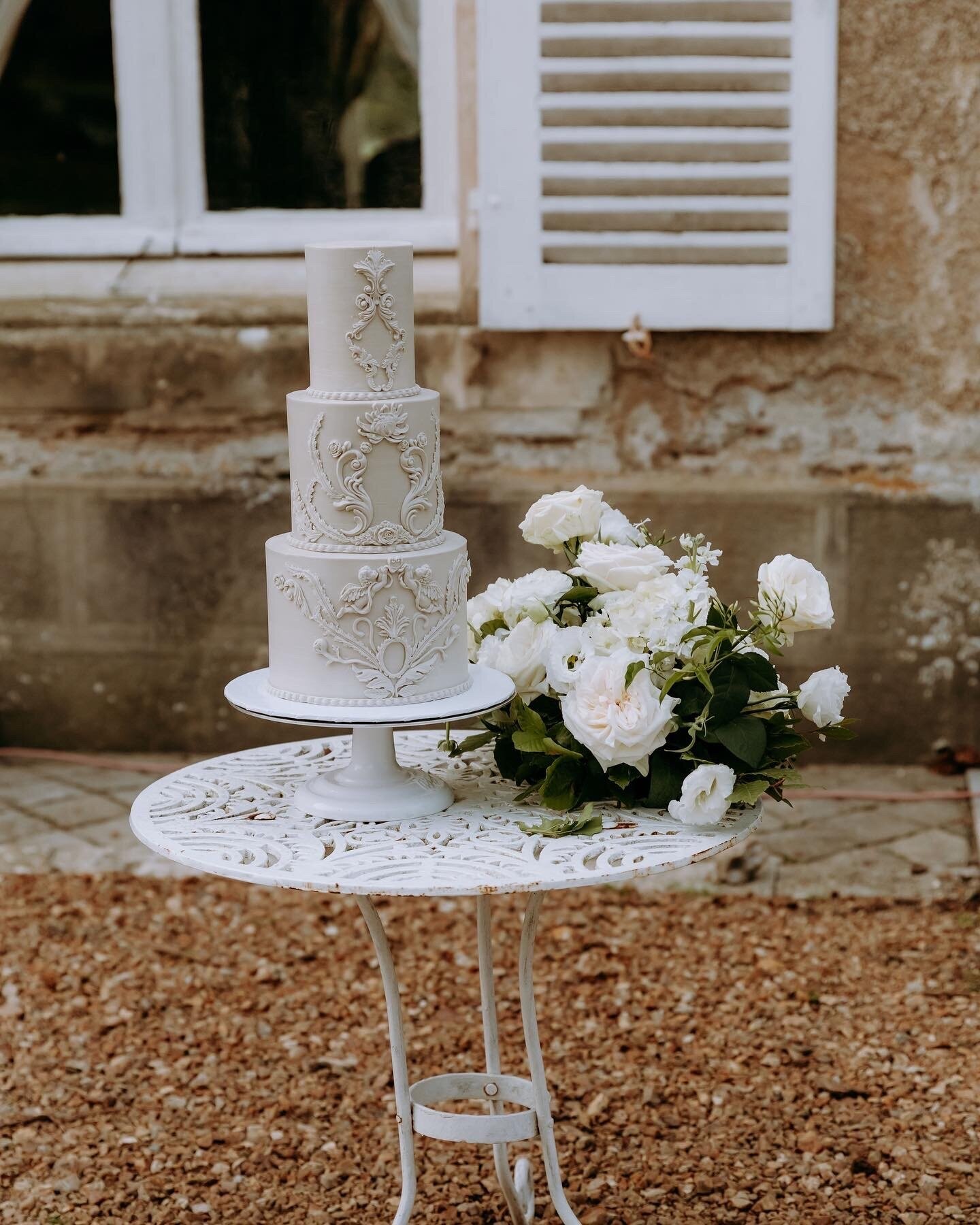 White two-tier wedding cake with lace piping on a garden table beside white roses at Château de Bouthonvilliers