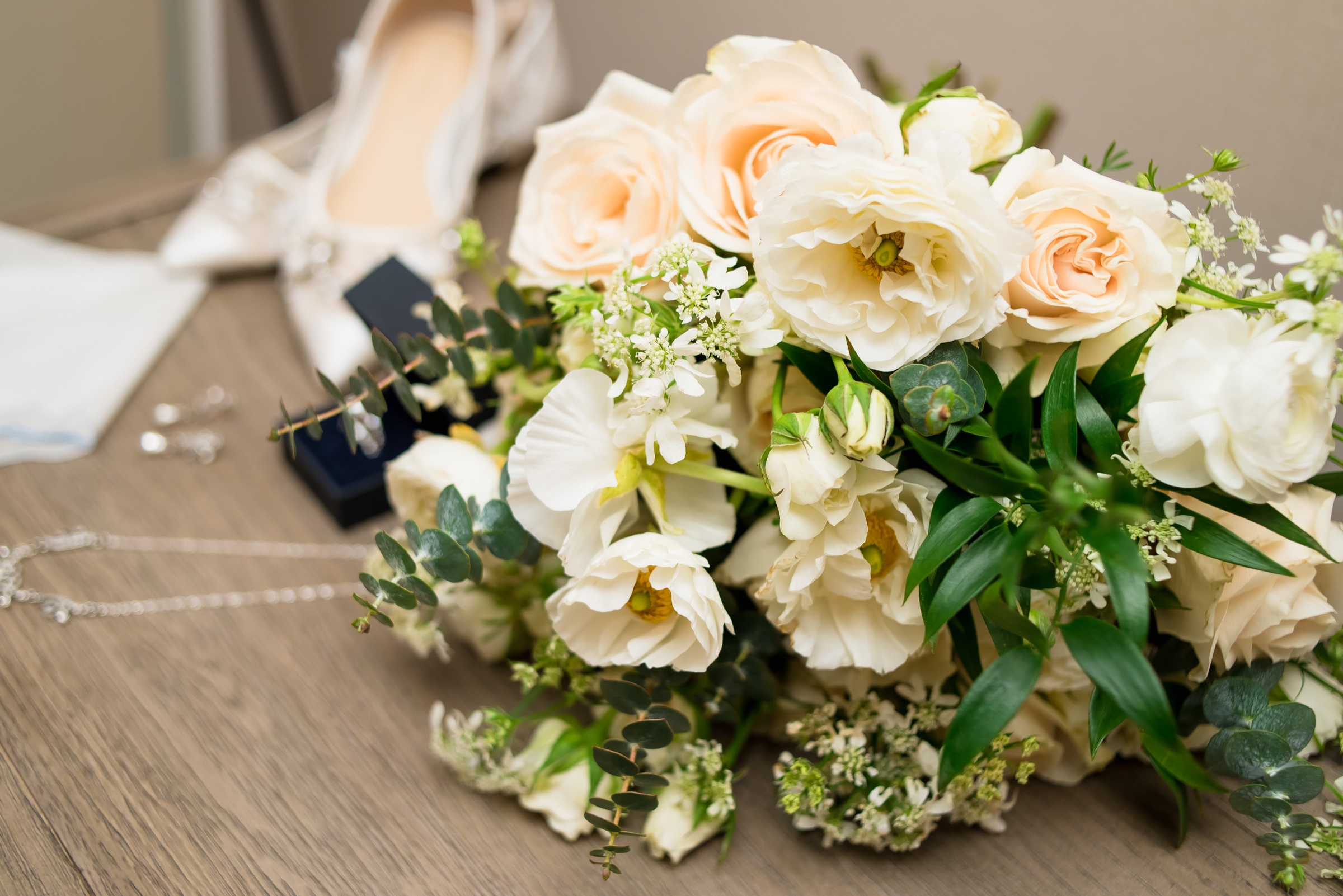 Bridal bouquet of peach garden roses, white ranunculus, lace flower, and eucalyptus with the bride's shoes and earrings
