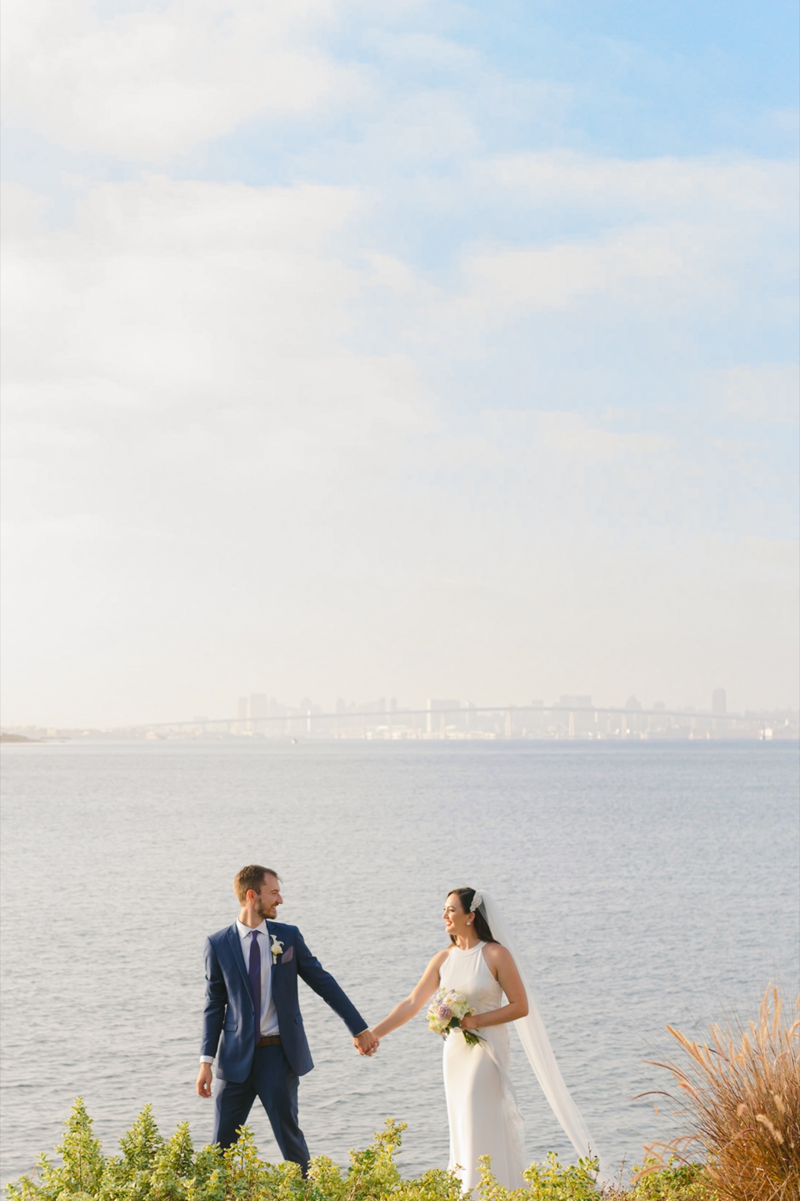 Julianne and David hand in hand at the water's edge at Loews Coronado Bay with the Coronado Bridge behind them