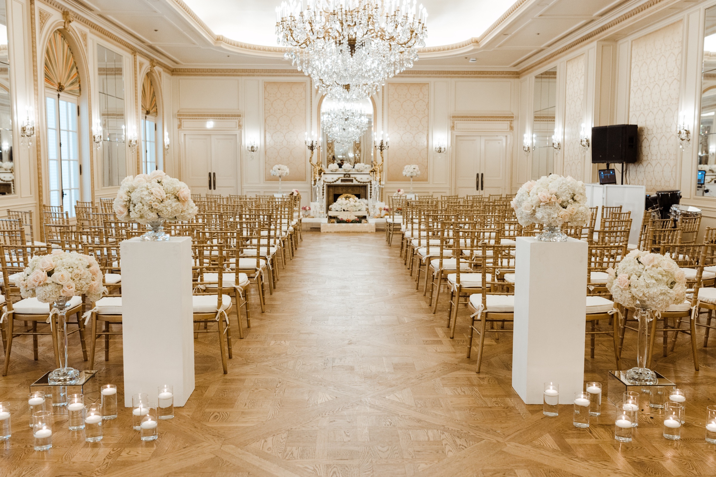 Ceremony room set with white floral pedestals and gold chairs under crystal chandeliers