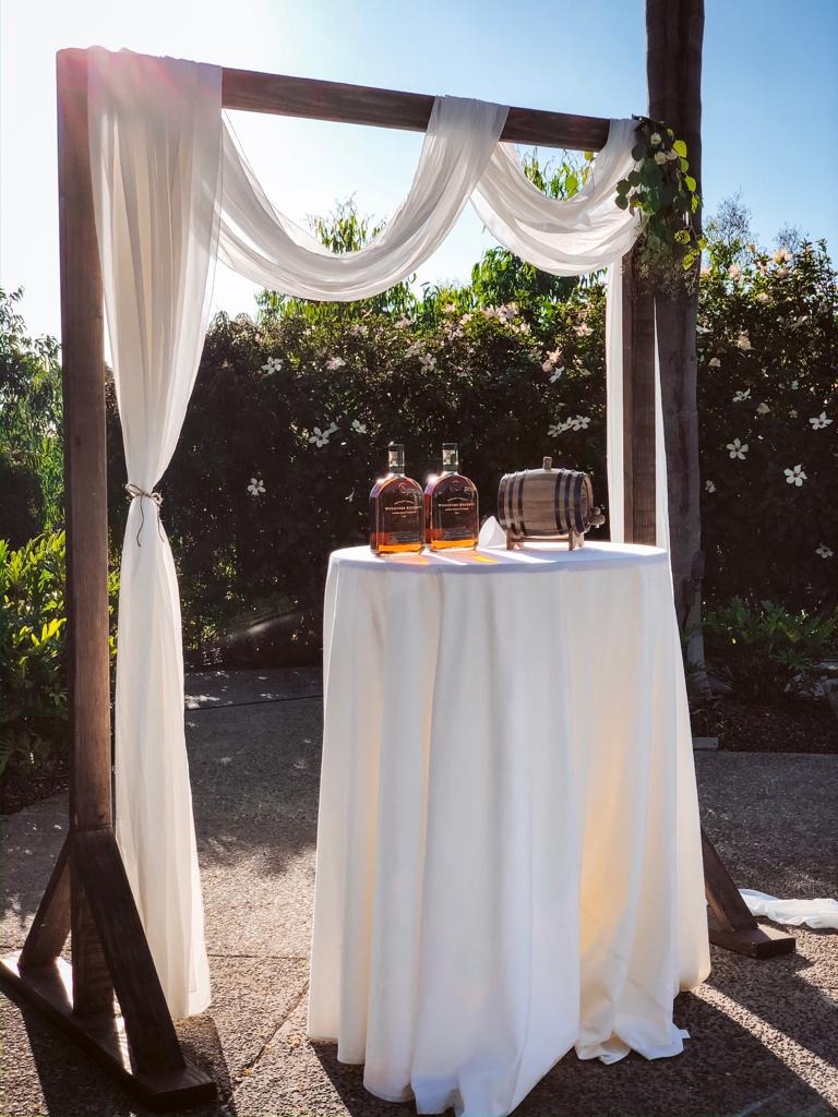 Whiskey bottles and a small oak barrel on a draped wooden arch table at Erica and Patrick’s Park Hyatt Aviara wedding